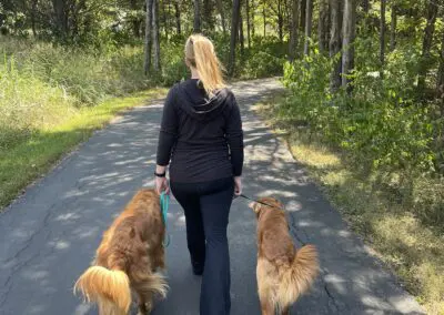 Woman walking two golden retrievers on shaded asphalt path through trees